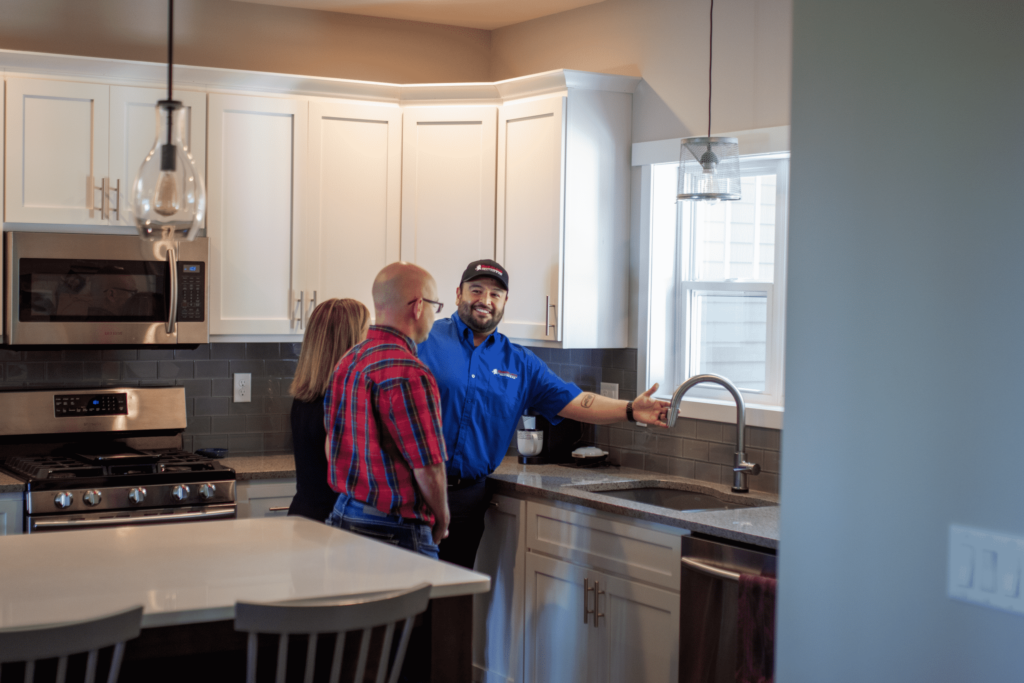 A person in a blue shirt is gesturing to a kitchen sink while talking to two other people. They stand in a modern kitchen with white cabinets and stainless steel appliances.
